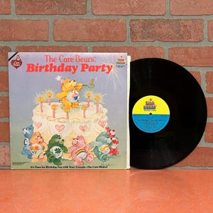 May include: A vinyl record and its square cardboard sleeve titled The Care Bears: Birthday Party. The sleeve features a yellow bear on a large cake surrounded by colorful Care Bears. The black vinyl record has a yellow and blue center label.