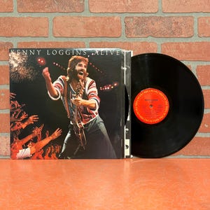 May include: A vinyl record album by Kenny Loggins titled "Alive." The album cover features a live performance photo of Loggins playing guitar, with the title and artist's name in white text. The record itself is black with a red and white label.