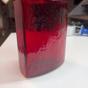 May include: A rectangular glass vase in a deep, translucent red hue. The exterior surface features a textured, hammered finish that creates variations in light reflection and color depth across the front and side panels of the vessel.