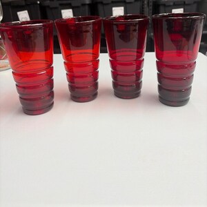 May include: A set of four matching ruby red drinking glasses. Each glass features a cylindrical shape with a series of horizontal, raised circular ridges or rings encircling the exterior body from the base up to the middle section.