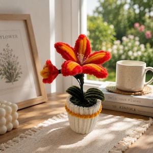Peut inclure: Une fleur artisanale en cure-pipe aux teintes rouge et orange vives, composée d'une grande fleur centrale et d'un bouton sur une tige sombre. Elle est plantée dans un pot blanc côtelé recouvert de tissu avec une bordure jaune, à la texture duveteuse.