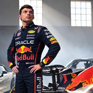May include: A dark navy blue racing suit featuring red side panels and multiple sponsor logos including Oracle, Red Bull, Honda, and Tag Heuer. The suit displays a yellow bull graphic on the chest and a small Dutch flag next to the name Max.