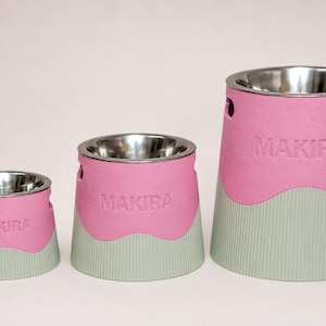 Pode incluir: Três tigelas para animais de estimação de alturas variadas, cada uma com um inserto de aço inoxidável e uma base de dois tons. A base é rosa no topo com uma borda ondulada e verde claro com nervuras verticais na parte inferior. A palavra MAKIRA está em relevo.