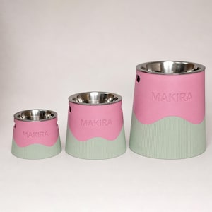 Puede incluir: Tres cuencos para mascotas de diferentes alturas, cada uno con un inserto de acero inoxidable, una sección superior rosa con el texto MAKIRA en relieve y una base acanalada de color verde claro con una transición ondulada.
