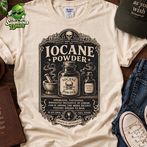 May include: A cream-colored t-shirt featuring a black graphic of a vintage apothecary label for Iocane Powder. The design includes a skull, mortar and pestle, and text reading "Princess Bride Apothecary," "Handle with Care," and "Death is Inevitable."