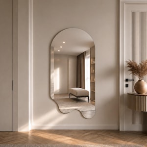 May include: A large, frameless wall mirror with an irregular, organic, wavy shape. The mirror features a smooth, reflective surface that captures the surrounding room, including a bench and curtains, and is mounted on a plain, light-colored wall.