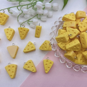 Peut inclure: Une collection de pendentifs en forme de fromage jaune avec des anneaux de saut argentés. Les breloques sont en forme de tranches de fromage et sont disposées sur une surface blanche avec un bol de breloques supplémentaires en arrière-plan.