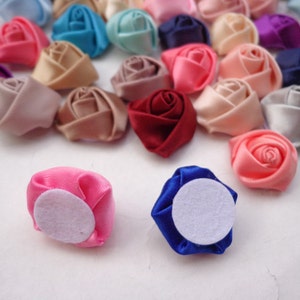 May include: Assortment of small fabric rose flower embellishments in various colors, including pink, red, blue, white, and beige. Some of the roses have a white felt backing.