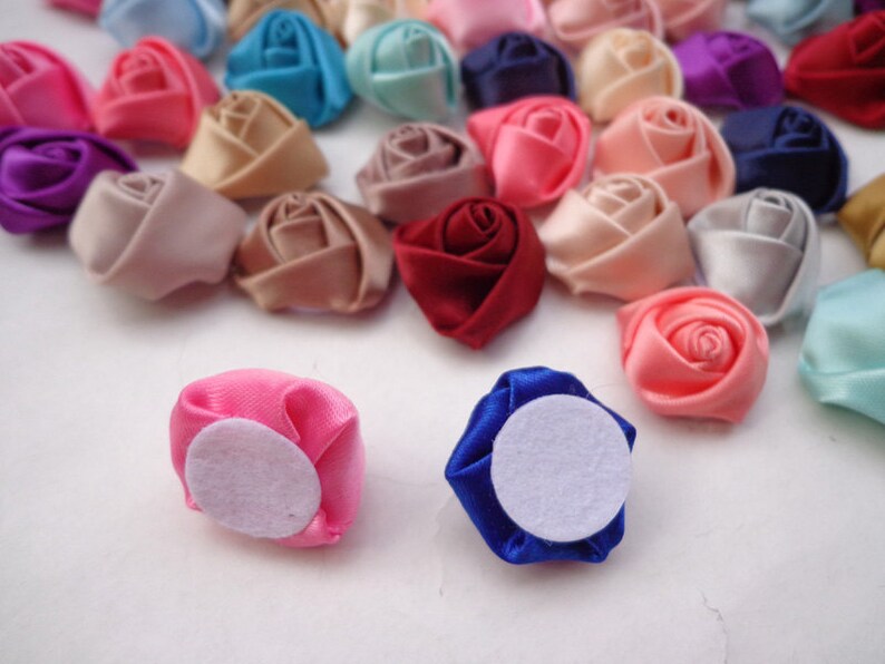 May include: Assortment of small fabric rose flower embellishments in various colors, including pink, red, blue, white, and beige. Some of the roses have a white felt backing.