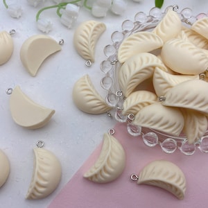May include: A collection of off-white, cream-colored,  dumpling-shaped charms with silver jump rings. The charms are arranged in a glass bowl and on a white surface.