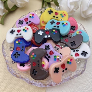 May include: A collection of colourful plastic game controller charms. The charms are in various colours, including blue, pink, purple, yellow, and white. Each charm features a heart, buttons, and a joystick.