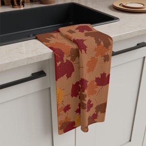 May include: A rectangular kitchen towel featuring a repeating pattern of autumn leaves in shades of deep red, burnt orange, golden yellow, and brown. The towel is draped over the edge of a white kitchen cabinet drawer beneath a black sink.
