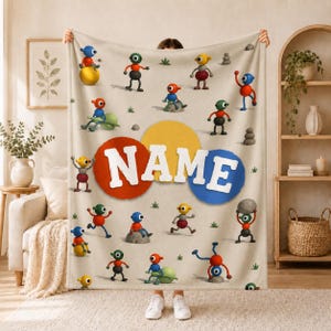 Pode incluir: Uma manta de lã bege com um padrão repetitivo de pequenas criaturas de desenho animado coloridas com múltiplos membros em vermelho, azul, amarelo e verde. O centro exibe a palavra NAME em letras brancas grandes sobre círculos sobrepostos.