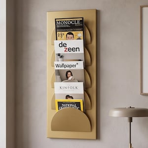 May include: A vertical gold metal wall-mounted magazine rack featuring five semi-circular pockets. The rack holds various magazines including Monocle, Dezeen, Wallpaper, Kinfolk, and National Geographic, displaying their covers in a stacked arrangement.