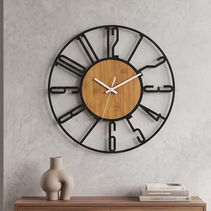 May include: A round wall clock featuring a black metal frame with large, stylised cut-out numbers and a central circular light brown bamboo wood face. The clock has three thin white hands positioned at the centre of the bamboo dial.