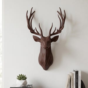 Puede incluir: Una cabeza de ciervo geométrica de color marrón oscuro para montaje en pared, con un diseño angular facetado y astas prominentes. La escultura está montada en una pared lisa, mostrando una forma estilizada de color sólido con bordes definidos y acabado mate.