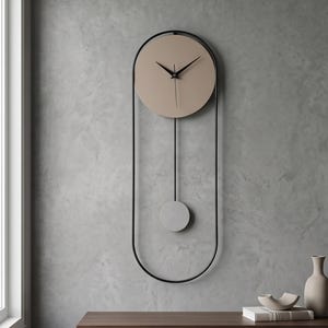 May include: A wall clock featuring a circular beige face with black hands, enclosed within a tall, thin black metal oval frame. A thin black rod extends downwards from the clock face to a small, circular light grey pendulum weight centred in the frame.