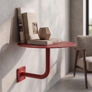 Puede incluir: Un estante de pared de metal rojo con una superficie superior circular y un brazo de soporte único unido a una placa base cuadrada. El estante sostiene dos libros con cubiertas beige y un pequeño objeto decorativo de madera de color claro.