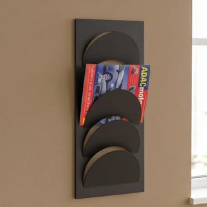 May include: A vertical black wall-mounted magazine rack featuring four semi-circular pockets. A magazine with a red cover and the text ADACmotorwelt is tucked into the second pocket from the top, displaying a blue car graphic on its front page.