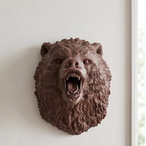 May include: A brown, sculpted wall-mounted bear head featuring a snarling expression with an open mouth, visible teeth, and detailed fur texture. The piece is rendered in a solid, monochromatic brown tone, capturing the animal's intense facial features.