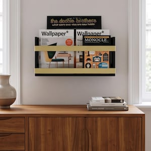 Puede incluir: Un revistero de pared con marco negro y dos barras metálicas horizontales de color dorado. El soporte contiene varias revistas, incluidos números de Wallpaper y Monocle, y una portada de álbum de los Doobie Brothers titulada What Were Once Vices.
