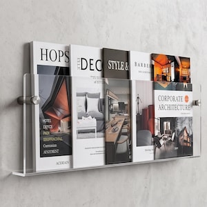 May include: A clear acrylic wall-mounted display rack holding several magazines and brochures. The rack features a transparent front panel secured by two silver-toned metal standoffs on either side, providing a floating appearance for the literature.