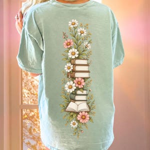 May include: A light sage green short-sleeve t-shirt featuring a vertical graphic on the back. The design displays a stack of five books intertwined with various colorful flowers, including white daisies and pink blossoms, with one open book at the base.