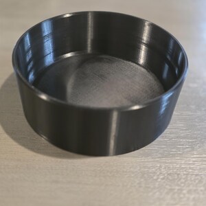 以下が含まれることがあります： 滑らかな内側と平らな底を持つ円形の黒い容器。この物体は、一定の厚みを持つ低い壁とマットな仕上げが特徴で、製造工程による繊細な水平線が外側に確認できます。
