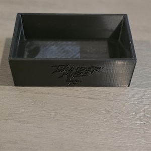 Könnte beinhalten: Ein rechteckiges schwarzes Tablett mit einem hohlen Zentrum und geraden Wänden. Die vordere Außenfläche zeigt den geprägten Text "THUNDER MIZER Exotics" in einer stilisierten Schriftart. Das Objekt hat eine matte Oberfläche mit sichtbaren 3D-Drucklinien.