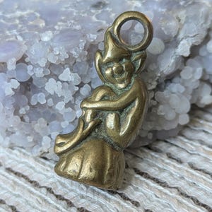 May include: A brass-coloured metal charm featuring a small, seated figure with pointed ears and a hat, depicted in a curled position. The charm includes a circular loop at the top for attachment, with a weathered, antique-style finish on the surface.