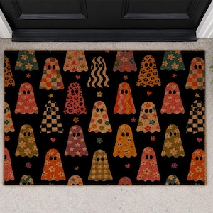 May include: A rectangular doormat with a black background featuring a repeating pattern of ghost shapes. Each ghost has a unique design including floral prints, leopard spots, plaid, and checkered patterns in shades of orange, brown, and red.