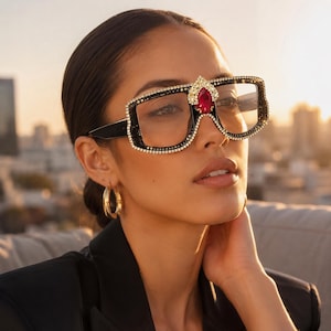 May include: Oversized square sunglasses featuring a black frame embellished with clear rhinestones along the edges. A prominent, teardrop-shaped red gemstone is set at the center of the bridge, framed by gold-tone metal accents.