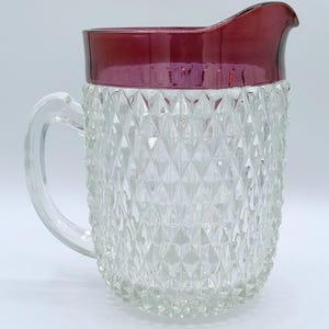 May include: A clear glass pitcher featuring a diamond-patterned texture on the body and a smooth, deep red rim. The pitcher includes a curved handle on the side and a pouring spout, showcasing a contrast between the textured clear glass and the solid red top.
