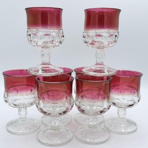 May include: A set of seven glass goblets featuring a clear base and stem with a textured, pressed pattern on the lower bowl. The upper portion of each goblet is finished with a deep magenta red gradient, creating a distinct contrast between the clear glass and the coloured rim.