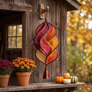 May include: A hanging spiral decoration crafted from crocheted yarn in a gradient of deep burgundy, vibrant orange, and soft yellow. The structure features an openwork pattern, a wooden bead at the top and bottom, and a multi-coloured yarn tassel.
