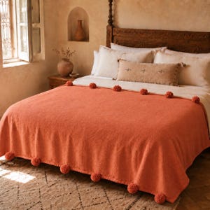 Puede incluir: Una colcha de color naranja quemado con una hilera de pompones a lo largo del borde superior y el dobladillo inferior. La cama está vestida con almohadas blancas y un cojín lumbar beige rectangular largo con pequeños detalles bordados.