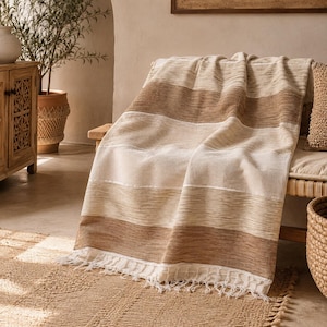Puede incluir: Una manta tejida con rayas horizontales en tonos crema, beige y marrón. La tela presenta un tejido texturizado visible y está rematada con un detalle de flecos a lo largo del borde inferior, creando una apariencia suave y superpuesta.