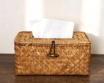 May include: A rectangular tissue box holder crafted from woven light brown natural fibres. The box features a hinged lid secured by a dark brown wooden toggle button attached to a thin black elastic cord, with white tissues protruding from the top.