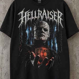 May include: A black short-sleeved t-shirt featuring a graphic print of the character Pinhead from Hellraiser. The design includes the text HELLRAISER in a stylised white font above the character holding a glowing red puzzle box cube.
