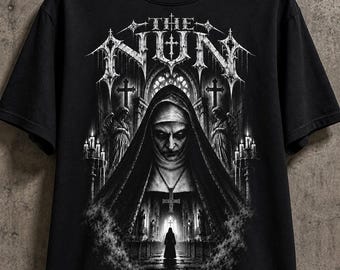Puede incluir: Una camiseta negra de manga corta con un estampado gráfico de una monja en una catedral oscura. El texto THE NUN está impreso en una fuente gótica en la parte superior. El diseño incluye velas, cruces y la silueta de una figura a lo lejos.