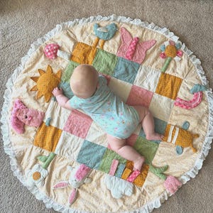 May include: A round, quilted play mat featuring a patchwork center in shades of green, pink, and yellow, surrounded by a cream-colored floral border with a ruffled edge. The mat includes various fabric appliques like a sun, butterfly, and flowers.