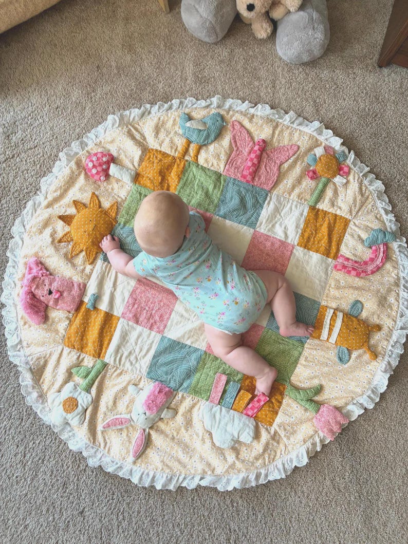 May include: A round, quilted play mat featuring a patchwork center in shades of green, pink, and yellow, surrounded by a cream-colored floral border with a ruffled edge. The mat includes various fabric appliques like a sun, butterfly, and flowers.