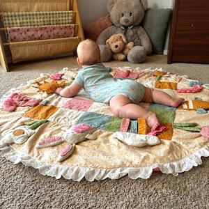 Op de afbeelding: Een ronde, gewatteerde speelmat voor baby's met een patchworkontwerp in tinten geel, groen en roze met bloemenpatronen. De mat bevat bevestigde pluche vormen zoals bloemen, een zon en een konijn, afgewerkt met een witte broderie anglaise rand.