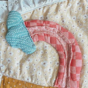 May include: A quilted fabric piece featuring a light yellow floral background with a white eyelet lace trim. A teal cloud with white dots and a coral pink checkered rainbow are appliqued onto the surface, alongside a mustard yellow star.