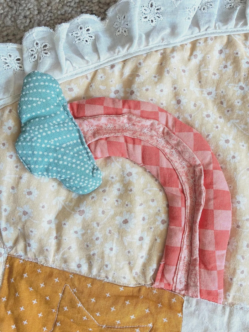 May include: A quilted fabric piece featuring a light yellow floral background with a white eyelet lace trim. A teal cloud with white dots and a coral pink checkered rainbow are appliqued onto the surface, alongside a mustard yellow star.