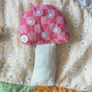 May include: A fabric mushroom applique featuring a pink and red checkered cap adorned with five clear plastic buttons. The mushroom has a plain off-white stem and is stitched onto a background of light yellow fabric with a white floral pattern.