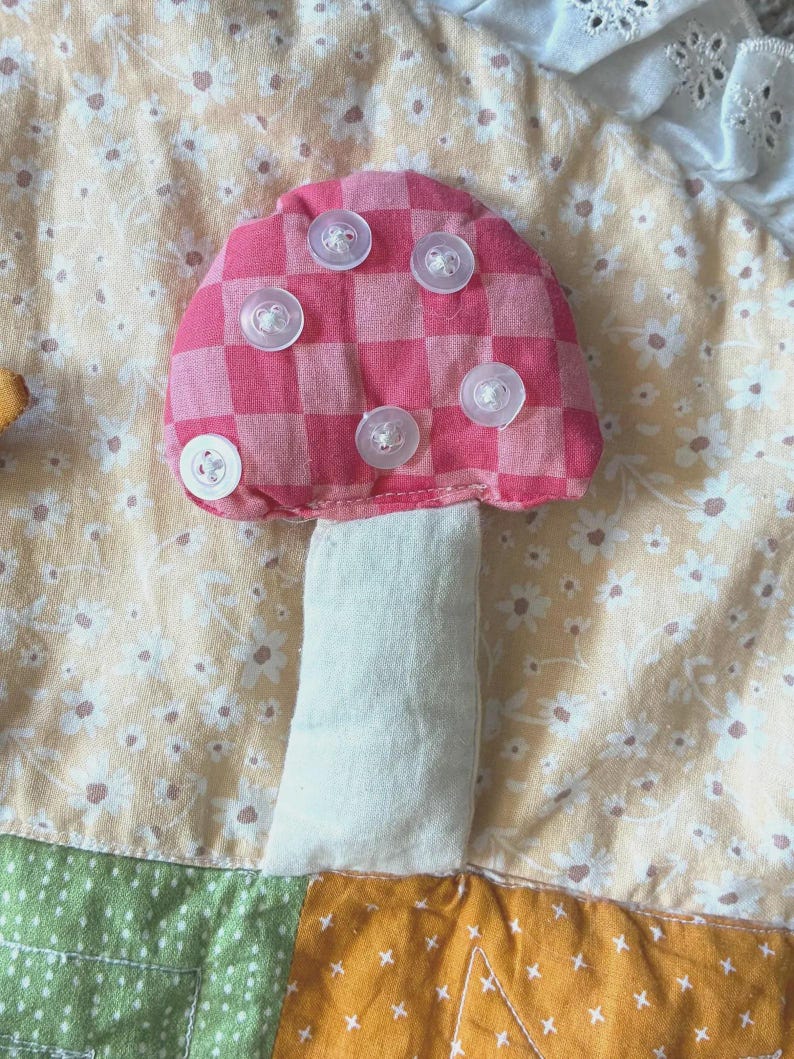 May include: A fabric mushroom applique featuring a pink and red checkered cap adorned with five clear plastic buttons. The mushroom has a plain off-white stem and is stitched onto a background of light yellow fabric with a white floral pattern.