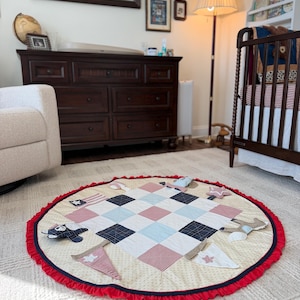 May include: A circular fabric play mat featuring a central patchwork design of red, white, and blue squares. The mat is bordered by a red ruffled edge and decorated with various small fabric appliques including stars, flags, and pennants.
