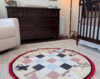 May include: A circular fabric play mat featuring a central patchwork design of red, white, and blue squares. The mat is bordered by a red ruffled edge and decorated with various small fabric appliques including stars, flags, and pennants.