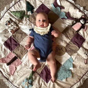 May include: A round, quilted play mat featuring a patchwork design in shades of cream, brown, and plum, accented with lace trim. The mat includes attached plush elements like an owl, trees, a whale, and a bear. A wooden disc reads six months.